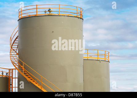 Gray cylindrical fuel tanks with yellow railings above blue cloudy sky Stock Photo
