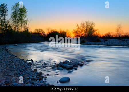 Blurred water of Boise River at sunset in Eagle with river rocks in foreground and bridge over river in background Stock Photo