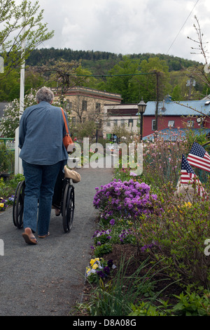 A woman pushes a wheelchair across Shelburne Falls Bridge of Flowers in Spring, with the Town of Buckland in the background. Stock Photo