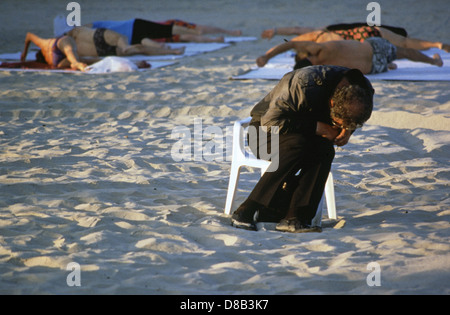 A homeless person sleeping on a chair with elderly people stretching at a sandy beach in Tel Aviv Israel Stock Photo