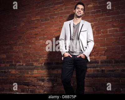 Portrait of a fashionable smiling young man wearing light gray blazer and black jeans standing at a brick wall Stock Photo