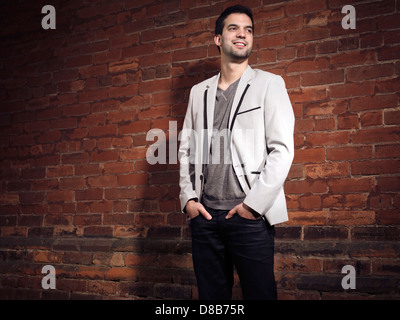 Artistic portrait of a smiling young man wearing light gray blazer and black jeans standing at a brick wall Stock Photo