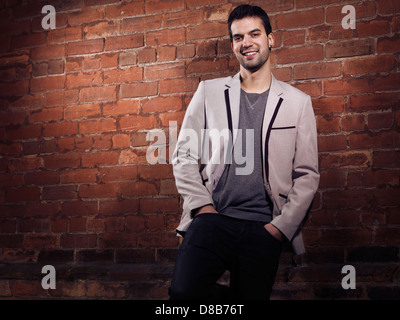 Artistic portrait of a smiling fashionably dressed young man wearing light gray blazer and black jeans standing at a brick wall Stock Photo