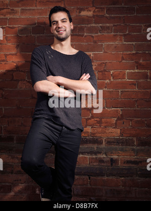 Expressive portrait of a smiling young man wearing black jeans and shirt standing at a brick wall Stock Photo