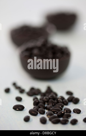 A small pile of black beans in front of three small bowls of black beans on a white wood surface. Stock Photo