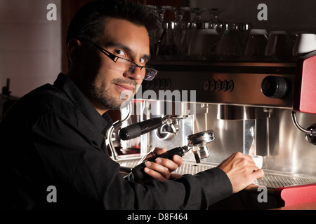 A male barista with short dark hair, a beard, and glasses is operating a professional espresso machine. He is wearing a black shirt and holding a portafilter, positioning it under the machine's group Stock Photo
