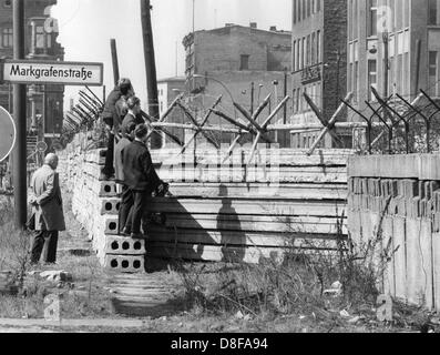 Westberliner Männer werfen am 17.04.1964 einen Blick über die im Bau befindliche vorverlegte Mauer an der Markgrafenstraße in Berlin. Am frühen Sonntagmorgen des 13. August 1961 wurde unter der Aufsicht von bewaffneten Streitkräften der DDR mit der Errichtung von Straßensperren aus Stacheldraht und dem Bau einer Mauer begonnen, um den Ostteil Berlins vom Westteil abzusperren. Die Mauer sollte den ständig steigenden Flüchtlingsstrom von Ost- nach West-Berlin stoppen. +++(c) dpa - Report+++ Stock Photo