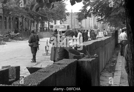 Construction workers of East Berlin extend the Berlin Wall from 'Potsdamer Platz' square in the direction of 'Lindenstrasse' street under the supervision of armed officers of the people's police in Berlin, GDR, 18 August 1961. The wall is build between two and five metres within the demarcation line of the sectors in East Berlin. Here, the borders of the sectors run along the house facades. Grit concrete slabs and hollow bricks, which were initially designed for the building of houses in East Berlin, are used for the construction. The construction workers are paid 1.28 Deutschmarks an hour. In Stock Photo