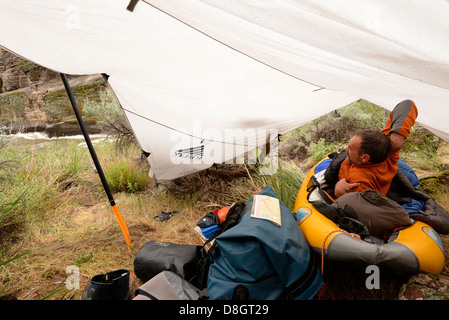 Kayaker waking from a night sleeping in inflatable kayak under a tarp on a trip down Idaho's Bruneau River. Stock Photo