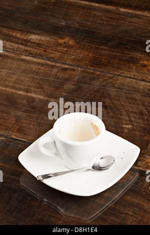 A white ceramic coffee cup, partially empty with coffee residue inside, sits on a matching white square saucer. A small metal spoon rests on the saucer beside the cup. The set is placed on a dark Stock Photo