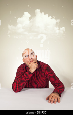 Worried young adult bald man sitting at the table with hand on his chin; cloud of questions above his head. Stock Photo