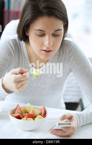 Portrait of a young woman eating fruit salad while browsing with her iphone Stock Photo