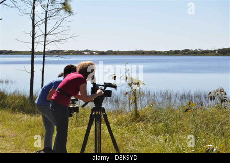 Two local broadcast journalists film a segment along the shore, with cameras set up to capture the coastal environment. The scene features the shoreline and calm waters as the backdrop for the news report. Stock Photo