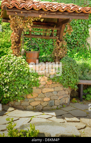 Old water well and a wooden bucket among ivy leaves in the garden Stock Photo