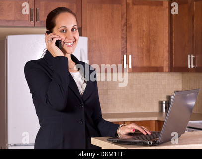 A business woman using a telephone and a laptop computer in her kitchen. Stock Photo