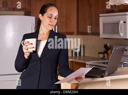 Business woman working on a laptop computer and drinking coffer in her kitchen. Stock Photo