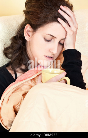 A young sick woman with a cup of tea covered with blanket at home, holding her head in pain Stock Photo