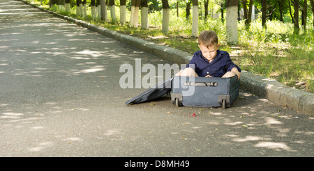 Little boy playing in a suitcase lying at the side of a tarred country road lined with leafy green trees Stock Photo