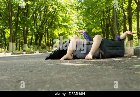 Small boy hidden in a suitcase with his legs protruding over the top as it lies on a rural road under shady trees Stock Photo