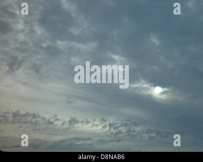 The sun is partially visible through dense cloud cover, creating a dramatic atmospheric effect. This type of weather condition is common during transitional periods in the atmosphere, often before or after a storm. The clouds diffuse the sunlight, creating a muted light effect on the landscape. The scene can evoke feelings of mystery and awe due to the unusual contrast of light and shadow. Stock Photo