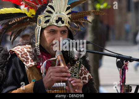 Manchester, UK. 1st June 2013. Sergio Fistely (MR) a street entertainer in Aztec Indian Native American costume and feather headdress in Manchester Piccadilly enjoying the varied attractions of the city centre with Peruvian buskers, costumed street muscians, playing musical accompaniment.  Pan flutes, panpipes, syrinx, flute are a group of musical instruments based on the principle of the closed tube, consisting of multiple pipes gradually increasing length.  Multiple varieties of panflutes have long been popular as folk instruments. The wooden pipes are typically made from bamboo, or cane. Stock Photo