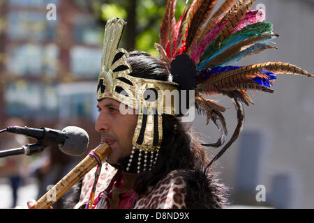 Manchester, UK. 1st June 2013. Sergio Fistely (MR) a street entertainer in Aztec Indian Native American costume and feather headdress in Manchester Piccadilly enjoying the varied attractions of the city centre with Peruvian buskers, costumed street muscians, playing musical accompaniment.  Pan flutes, panpipes, syrinx, flute are a group of musical instruments based on the principle of the closed tube, consisting of multiple pipes gradually increasing length.  Multiple varieties of panflutes have long been popular as folk instruments. The wooden pipes are typically made from bamboo, or cane. Stock Photo