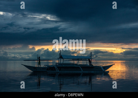 Boy standing on bow of outrigger bangka - a traditional Filipino fishing boat - in sunset with long pole in his hands Stock Photo