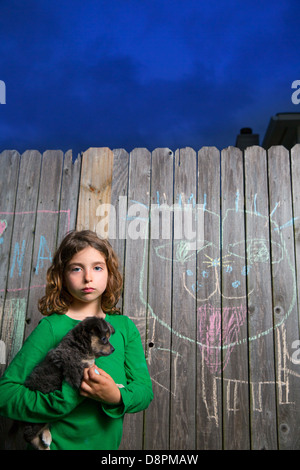 children girl holding puppy dog after chalk painting on backyard wood fence Stock Photo