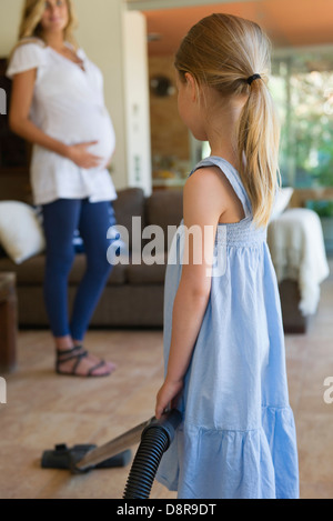 Little girl vacuuming, pregnant mother in background Stock Photo