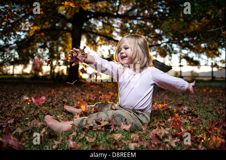 Friendly toddler sitting on a blanket of autumn leafs with the setting sun behind a big tree. Stock Photo