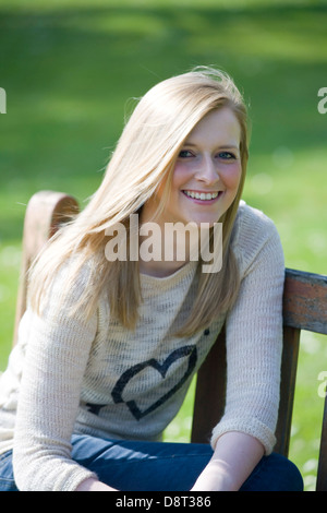 Pretty blonde girl siting on a park bench looking towards camera. Stock Photo