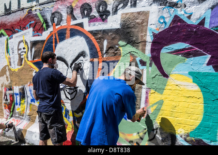 Youngsters with spray cans in alley spraying colourful graffiti on wall of building in town Stock Photo