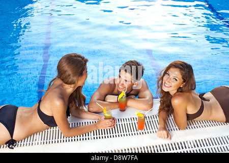 Handsome young man standing in a sparkling blue swimming pool chatting to two beautiful women in bikinis sunbathing on the edge. Stock Photo