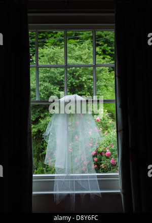 A wedding veil hung from a window overlooking a green garden. Stock Photo