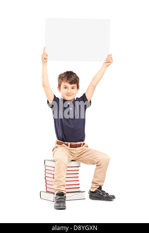 Happy school boy holding a blank panel above his head, seated on a pile of books isolated on white background Stock Photo