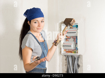 Attractive young female electrician standing in front of an open fuse box with a wooden mallet in one hand and a screwdriver in Stock Photo
