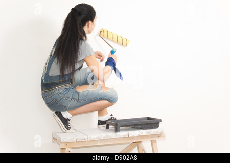 Young woman painting a wall with a roller balanced on a trestle or wooden table for extra height Stock Photo