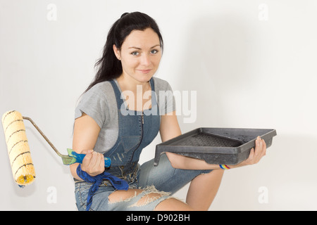 Pretty young woman with a paint tray and roller in her hands crouching down as she decorates the walls of her home Stock Photo