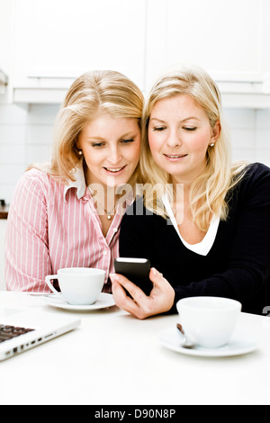 Two young women using mobile phone in kitchen Stock Photo