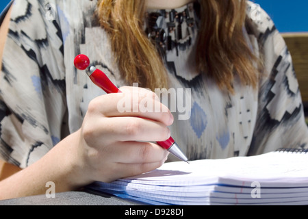 Medium shot of young female student, sitting outside studying/writing on a notebook paper with a red pen. Stock Photo