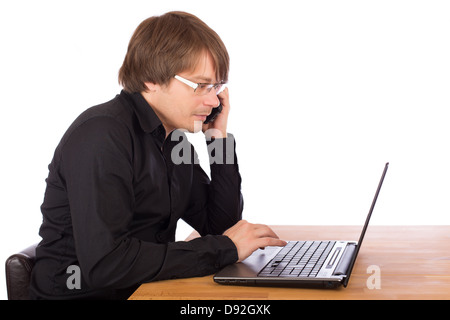 Young business man with black shirt and mobile phone working seriously on a laptop. Isolated on white background. Stock Photo
