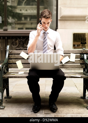 Businessman using a smart phone while using a laptop and sitting on bench with sticky notes Stock Photo