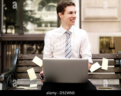 Smiling businessman holding smart phone while using a laptop sitting on bench with sticky notes Stock Photo