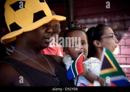 A father wearing novelty football hat dog tag holds daugther who waves South African flag while they watch live concert as part Stock Photo