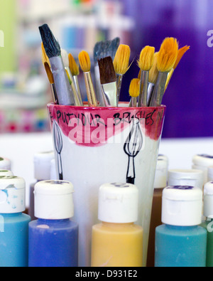 Kids art brushes peeping from a home made ceramic jar, surrounded by colourful  paints in front of a colourful studio background Stock Photo