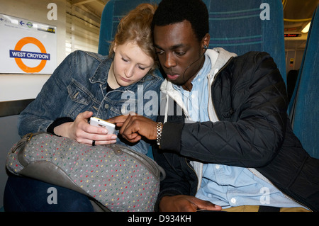 Interracial teenage couple using mobile phone, seated on train at West Croydon National Rail station and Tramlink stop in South London, England, UK. Stock Photo