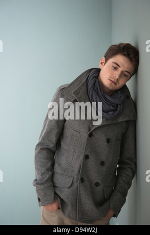 Portrait of Young Man wearing Grey Scarf and Jacket, Leaning against Wall, Studio Shot on Grey Background Stock Photo