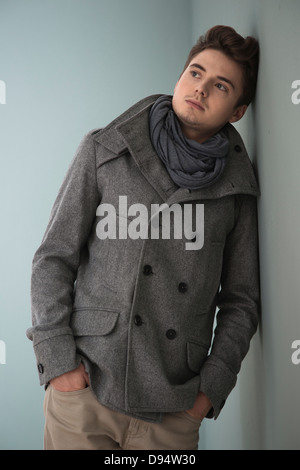 Portrait of Young Man wearing Grey Scarf and Jacket, Leaning against Wall, Studio Shot on Grey Background Stock Photo