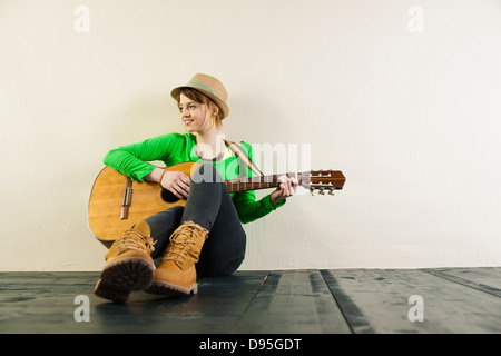 Portrait of Teenage Girl Sitting on Floor, Wearing Hat and Playing Acoustic Guitar, Studio Shot on White Background Stock Photo