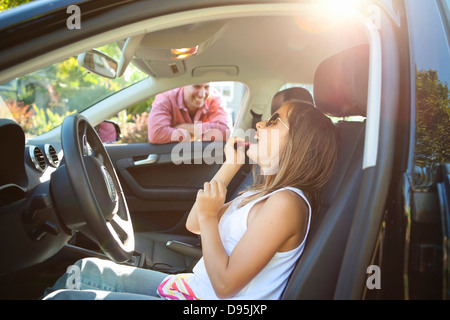 Young girl applying Lip Gloss pretending be old enough drive as her smiling father watches on on sunny summer evening in Stock Photo